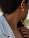 A close-up side profile of a woman wearing a small gold V-shaped stud earring, with her dark hair and white blazer visible against a softly blurred background. ACHIEVE TRIANGLE GEOMETRIC HUGGIE EARRINGS IN 18K GOLD Over Sterling Silver by Sonia Hou Jewelry