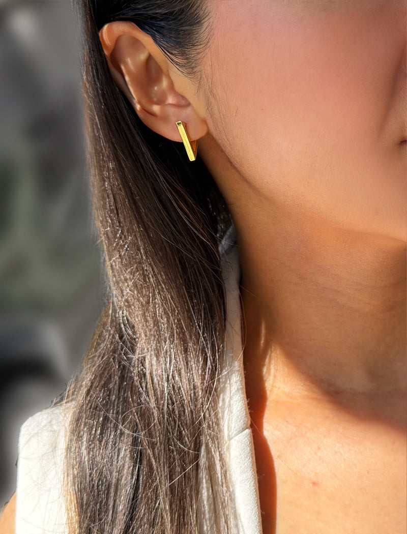 A close-up of a woman's ear wearing a small gold geometric stud earring, with her hair and shoulder visible against a softly blurred background. ACHIEVE TRIANGLE GEOMETRIC HUGGIE EARRINGS IN 18K GOLD Over Sterling Silver by Sonia Hou Jewelry