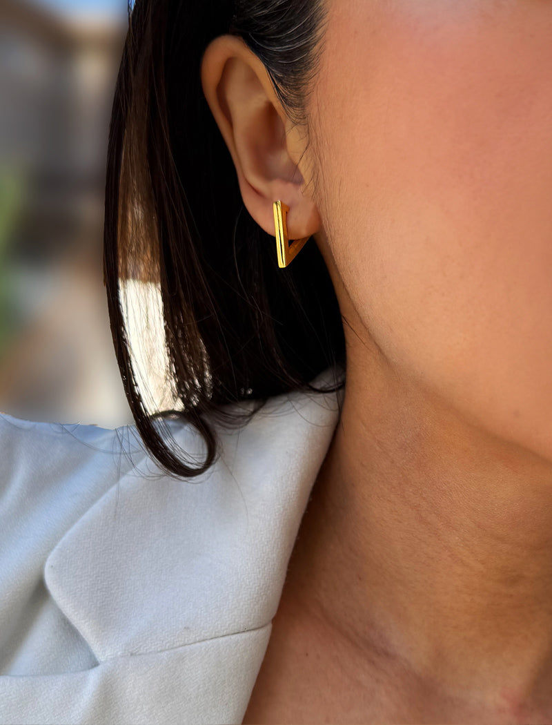 A close-up of a person's ear wearing a small gold hoop earring, with dark hair and a light-colored top visible against a softly blurred background. ACHIEVE TRIANGLE GEOMETRIC HUGGIE EARRINGS IN 18K GOLD Over Sterling Silver by Sonia Hou Jewelry