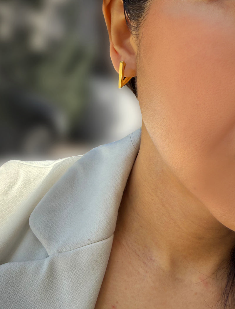 A close-up of a person's ear wearing a small geometric gold hoop earring, with part of a light-colored blazer visible against a softly blurred outdoor background. ACHIEVE TRIANGLE GEOMETRIC HUGGIE EARRINGS IN 18K GOLD Over Sterling Silver by Sonia Hou Jewelry