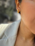 A close-up of a person's ear wearing a small geometric gold hoop earring, with part of a light-colored blazer visible against a softly blurred outdoor background. ACHIEVE TRIANGLE GEOMETRIC HUGGIE EARRINGS IN 18K GOLD Over Sterling Silver by Sonia Hou Jewelry