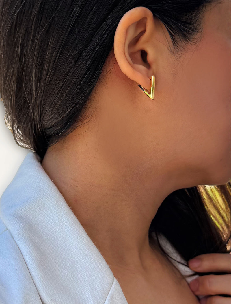 A close-up of a person's ear wearing a small gold chevron-shaped stud earring, with dark hair and a light blue collared shirt visible. ACHIEVE TRIANGLE GEOMETRIC HUGGIE EARRINGS IN 18K GOLD Over Sterling Silver by Sonia Hou Jewelry