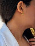 A close-up of a person's ear wearing a small gold chevron-shaped stud earring, with dark hair and a light blue collared shirt visible. ACHIEVE TRIANGLE GEOMETRIC HUGGIE EARRINGS IN 18K GOLD Over Sterling Silver by Sonia Hou Jewelry
