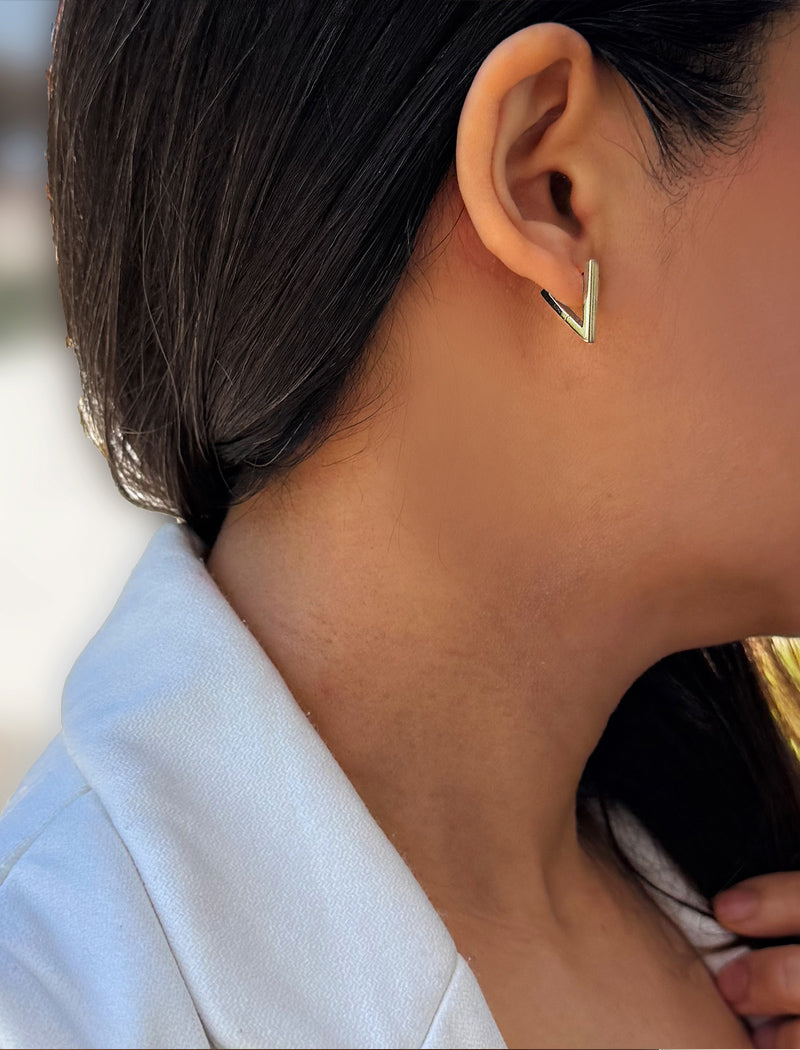 A close-up of a person's ear wearing a small gold chevron-shaped stud earring, with dark hair and a light blue collared shirt visible. ACHIEVE TRIANGLE GEOMETRIC HUGGIE EARRINGS IN  Sterling Silver by Sonia Hou Jewelry
