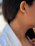 A close-up of a person's ear wearing a small gold chevron-shaped stud earring, with dark hair and a light blue collared shirt visible. ACHIEVE TRIANGLE GEOMETRIC HUGGIE EARRINGS IN  Sterling Silver by Sonia Hou Jewelry