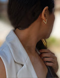 A close-up side profile of a woman wearing a small gold V-shaped stud earring, with her hand near her collarbone and a white sleeveless top visible against a softly blurred outdoor background. ACHIEVE TRIANGLE GEOMETRIC HUGGIE EARRINGS IN  Sterling Silver by Sonia Hou Jewelry