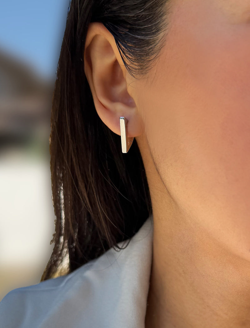 A close-up of a woman's ear wearing a minimalist, polished silver vertical bar stud earring. ACHIEVE TRIANGLE GEOMETRIC HUGGIE EARRINGS IN  Sterling Silver by Sonia Hou Jewelry