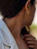 A close-up side profile of a woman wearing a small gold V-shaped stud earring, with her dark hair and white top visible against a softly blurred background. ACHIEVE TRIANGLE GEOMETRIC HUGGIE EARRINGS IN  Sterling Silver by Sonia Hou Jewelry