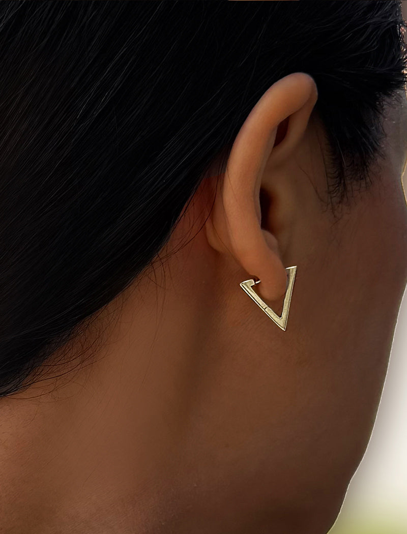 A close-up side profile of a person wearing a small gold triangular hoop earring. ACHIEVE TRIANGLE GEOMETRIC HUGGIE EARRINGS IN  Sterling Silver by Sonia Hou Jewelry