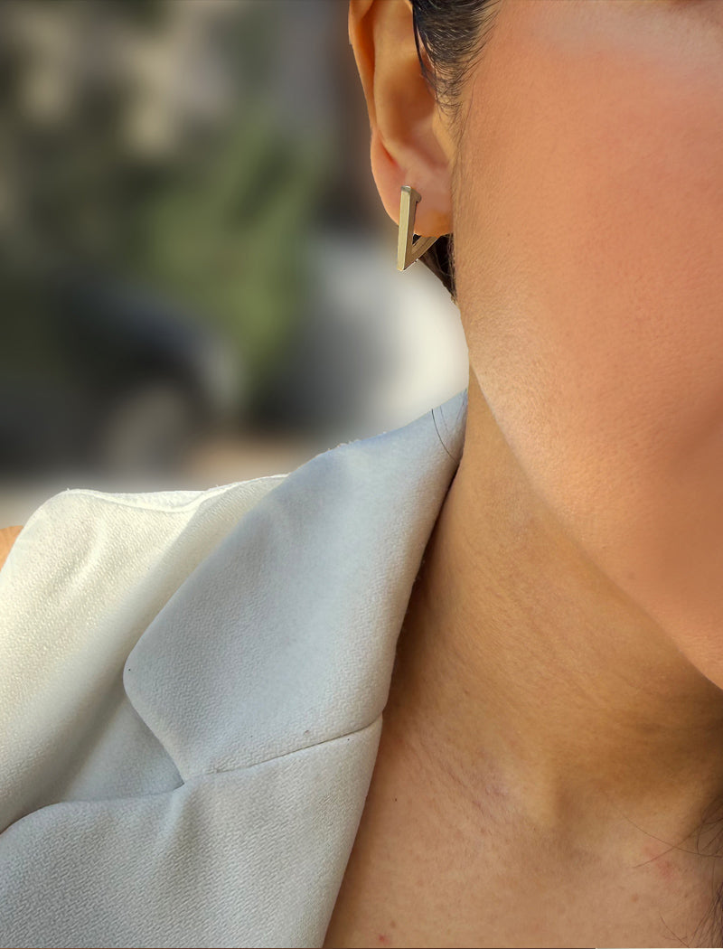 A close-up lifestyle shot of a model wearing a small, geometric gold-toned stud earring. The earring features a triangular prong design. ACHIEVE TRIANGLE GEOMETRIC HUGGIE EARRINGS IN  Sterling Silver by Sonia Hou Jewelry