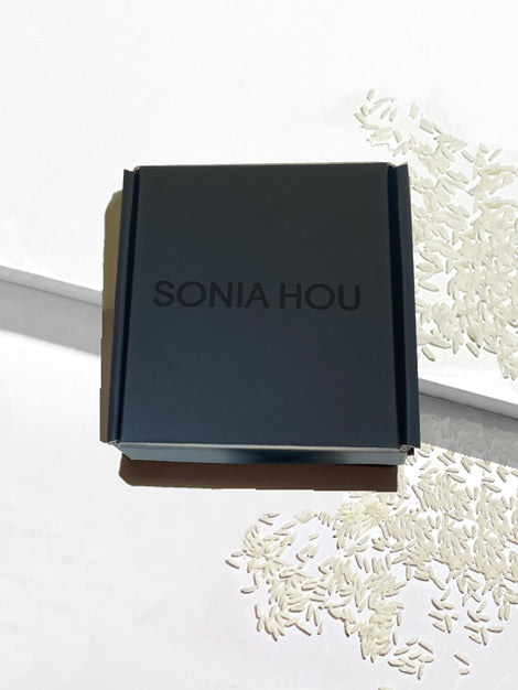 A dark charcoal gray square product box with the brand name 'SONIA HOU' printed in a matching dark gray font on the lid, styled on a white surface with scattered white grains.
