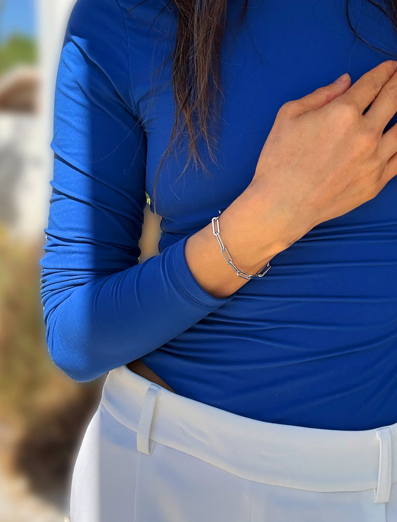 A close-up of a person wearing a bright royal blue long-sleeve athletic top and white bottoms, with a stainless steel silver chain bracelet visible on the wrist. Designed by AAPI celebrity jewelry designer SONIA HOU.