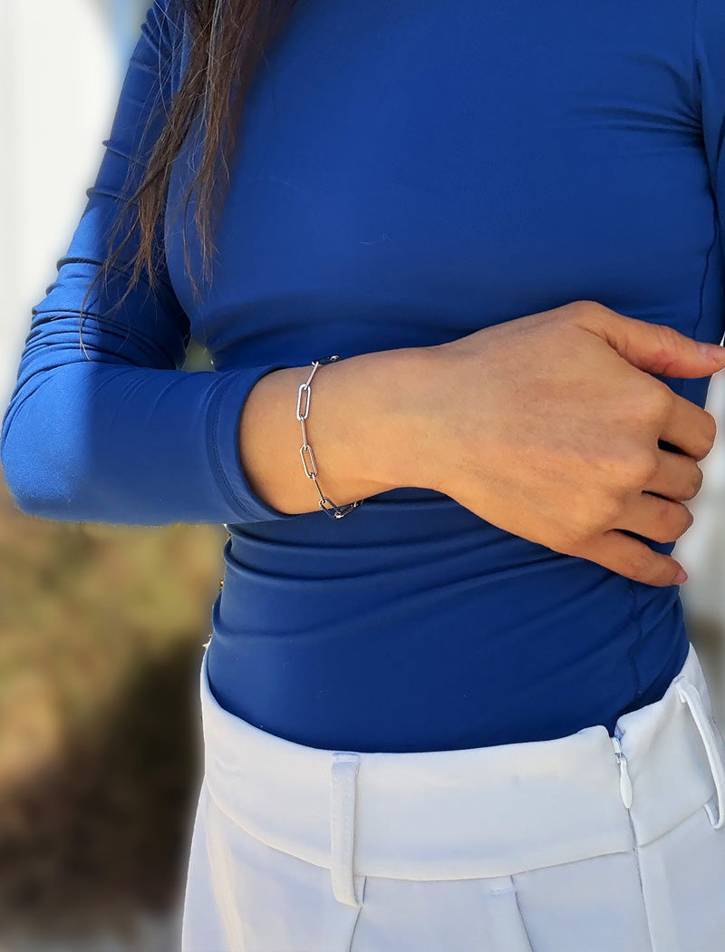 A close-up of a person wearing a stainless silver paperclip chain bracelet on their left wrist, paired with a royal blue long-sleeve top and white pants. Designed by AAPI celebrity jewelry designer SONIA HOU.