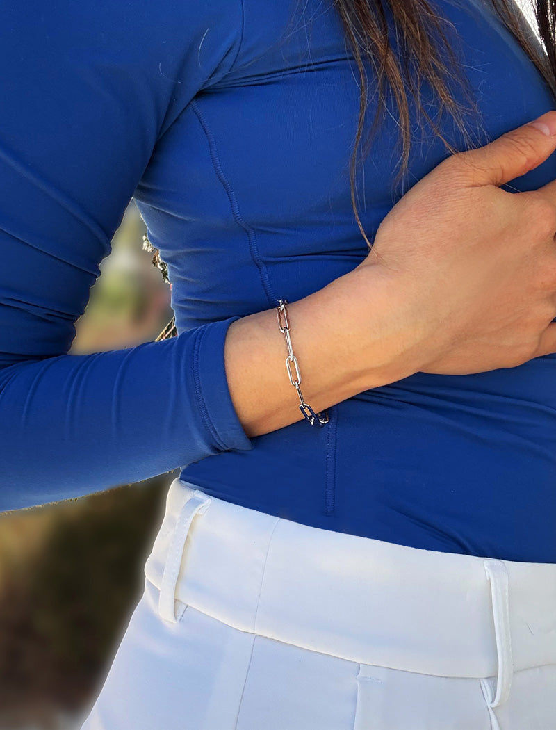 A close-up of a person wearing a stainless steel silver chain link bracelet on their wrist, dressed in a royal blue long-sleeve top and white pants. Designed by AAPI celebrity jewelry designer SONIA HOU.