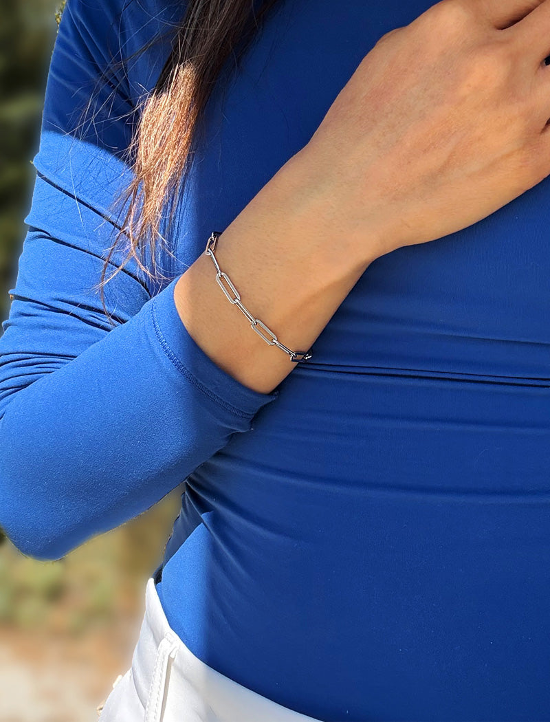 A close-up of a person wearing a stainless silver paperclip chain bracelet on their wrist, paired with a blue long-sleeve top and white pants. Designed by AAPI celebrity jewelry designer SONIA HOU.