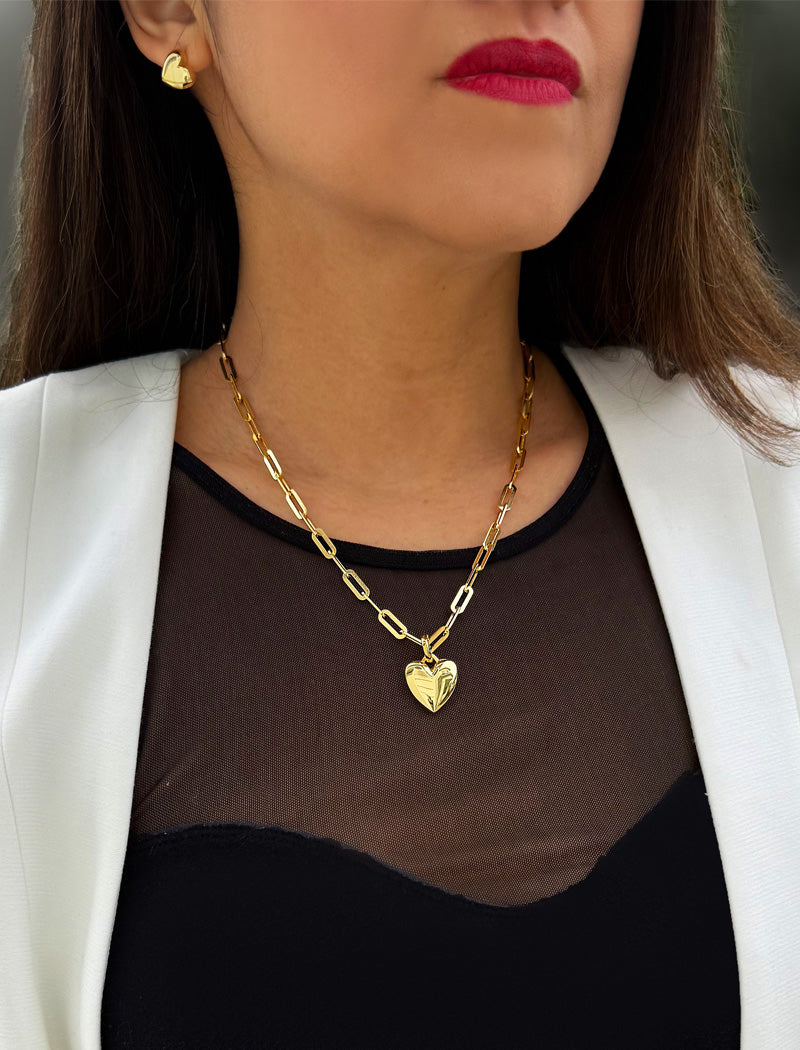 Woman wearing a gold chain necklace with a heart pendant, white blazer, and red lipstick. 18K Gold vermeil chain necklace with a heart pendant worn by a person. Featuring a bold yet feminine Puffy Heart charm suspended from a sleek link chain, this bracelet radiates effortless confidence and charm. Designed to symbolize love, self-expression, and empowerment, the puffy heart serves as a daily reminder to lead with compassion and strength — Designed by AAPI celebrity jewelry designer SONIA HOU.
