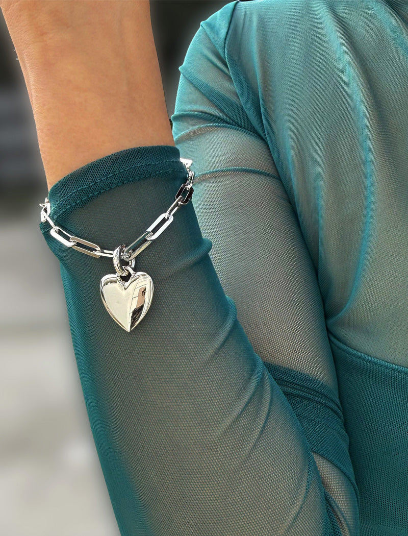 Sterling Silver bracelet with a heart-shaped pendant on a person wearing a teal top. Featuring a bold yet feminine Puffy Heart charm suspended from a sleek link chain, this bracelet radiates effortless confidence and charm. Designed to symbolize love, self-expression, and empowerment, the puffy heart serves as a daily reminder to lead with compassion and strength — for yourself and others. Designed by AAPI celebrity jewelry designer SONIA HOU.
