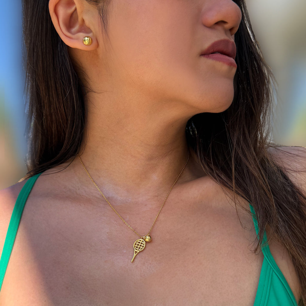A close-up lifestyle shot of a woman wearing a delicate gold tennis racket and ball pendant necklace and a matching gold tennis ball stud earring, set against a soft, blurred outdoor background. Designed by Sonia Hou, a celebrity AAPI inclusive demi-fine jewelry designer. Available in 18K Gold Over Sterling Silver, Sterling Silver and 18K Gold PVD Sterling Silver (Waterproof).