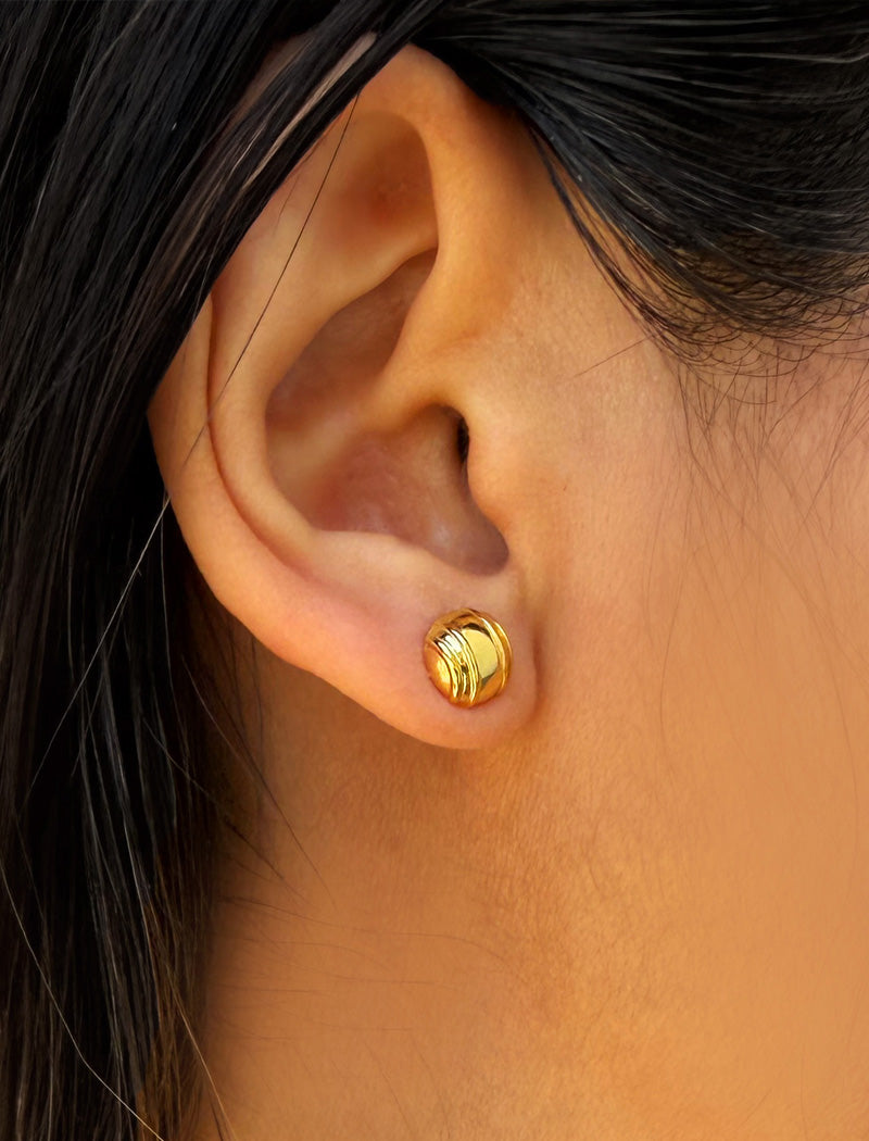 A close-up of a person's ear wearing a small gold tennis ball stud earring with a textured, ridged circular face, designed by Sonia Hou, a celebrity AAPI inclusive demi-fine jewelry designer. Available in 18K Gold Over Sterling Silver, Sterling Silver and 18K Gold PVD Stainless Steel (Waterproof).