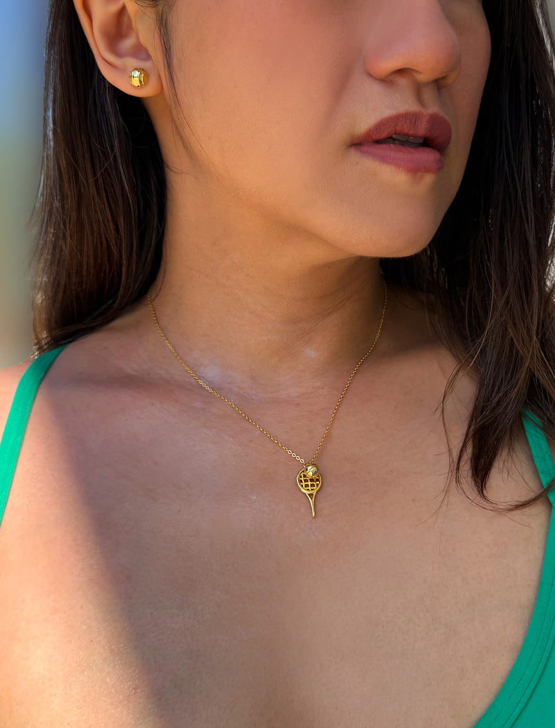 A close-up lifestyle shot of a woman wearing a delicate gold chain tennis ball and racquet necklace featuring a grid pattern, paired with a matching gold stud tennis ball earring. Designed by Sonia Hou, a celebrity AAPI inclusive demi-fine jewelry designer. Available in 18K Gold Over Sterling Silver, Sterling Silver and 18K Gold PVD Stainless Steel (Waterproof).