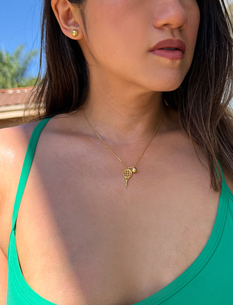 A close-up lifestyle shot of a woman wearing a delicate gold tennis ball & racquet pendant necklace and a matching gold stud tennis ball earring earring, set against a bright outdoor background. Designed by Sonia Hou, a celebrity AAPI inclusive demi-fine jewelry designer. Available in 18K Gold Over Sterling Silver, Sterling Silver and 18K Gold PVD Stainless Steel (Waterproof).