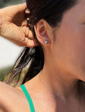 A close-up of a woman's ear wearing a small round tennis ball stud earring with a clear faceted gemstone in a silver-toned setting. Her hand is adjusting her hair, and she is wearing a green strap and a delicate silver necklace. Designed by Sonia Hou, a celebrity AAPI inclusive demi-fine jewelry designer. Available in 18K Gold Over Sterling Silver, Sterling Silver and 18K Gold PVD Stainless Steel (Waterproof).