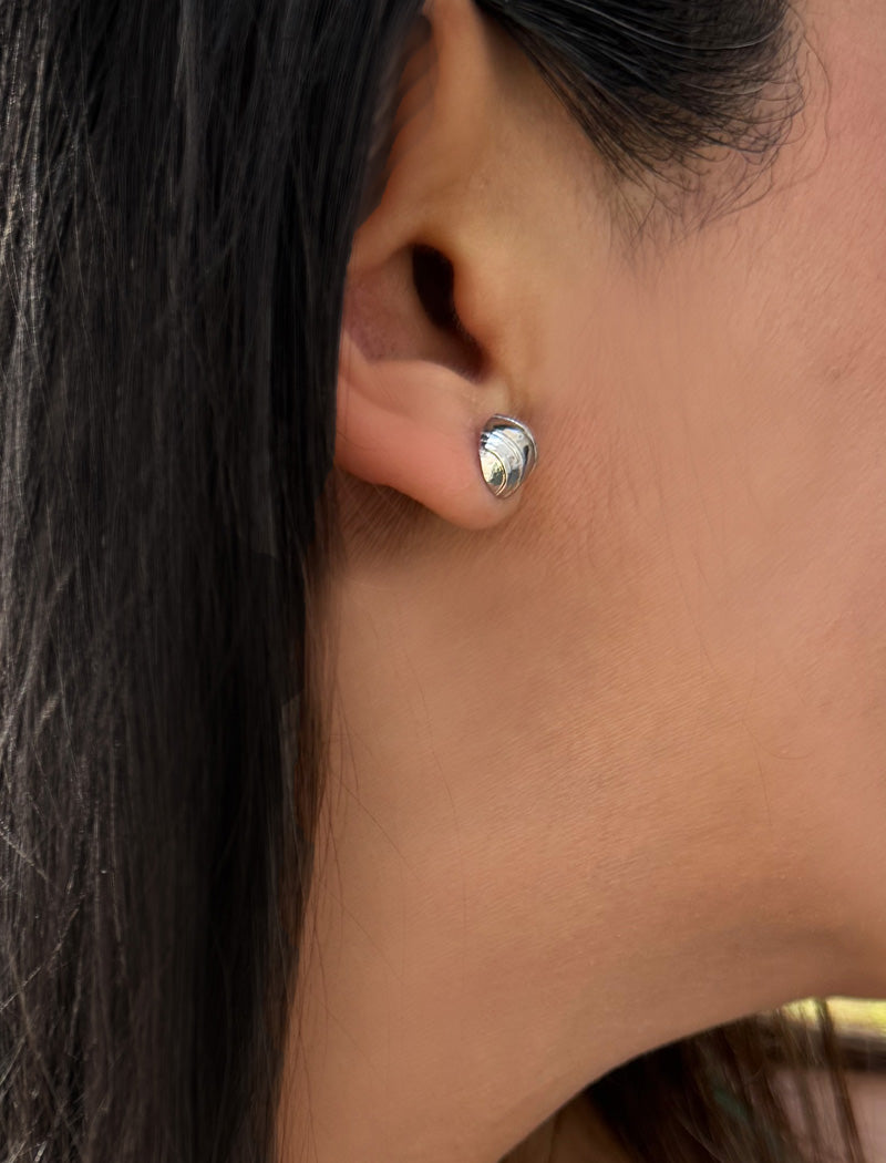 A close-up of a person's ear wearing a small, polished silver tennis ball stud earring with a textured, shell-like surface. Designed by Sonia Hou, a celebrity AAPI inclusive demi-fine jewelry designer. Available in 18K Gold Over Sterling Silver, Sterling Silver and 18K Gold PVD Stainless Steel (Waterproof).
