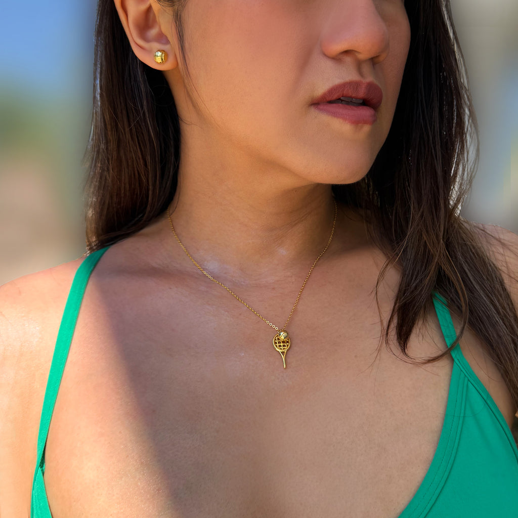 A close-up lifestyle shot of a woman wearing a delicate gold tennis racquet and ball pendant necklace and a matching gold tennis ball stud earring. The pendant features a stylized sun or flower design. Designed by Sonia Hou, a celebrity AAPI inclusive demi-fine jewelry designer. Available in 18K Gold Over Sterling Silver, Sterling Silver and 18K Gold PVD Sterling Silver (Waterproof).