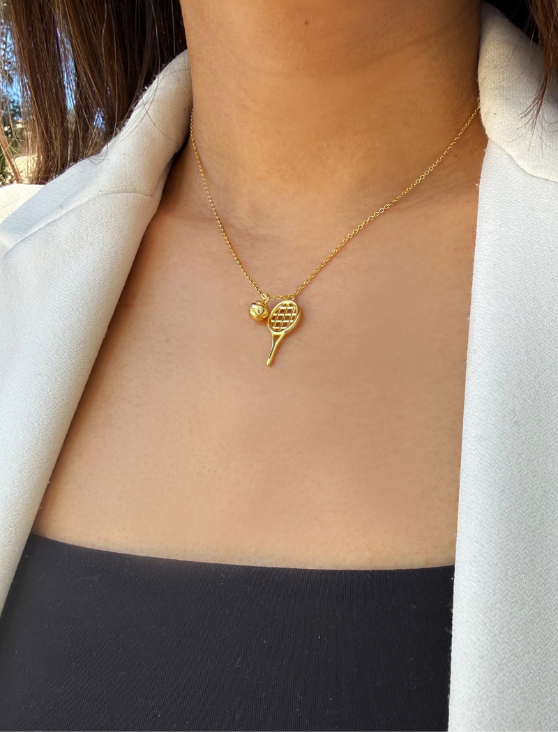 A close-up of a model wearing a delicate gold necklace with a tennis racket and tennis ball pendant, styled with a white blazer and black top. Designed by Sonia Hou, a celebrity AAPI inclusive demi-fine jewelry designer. Available in 18K Gold Over Sterling Silver, Sterling Silver and 18K Gold PVD Sterling Silver.