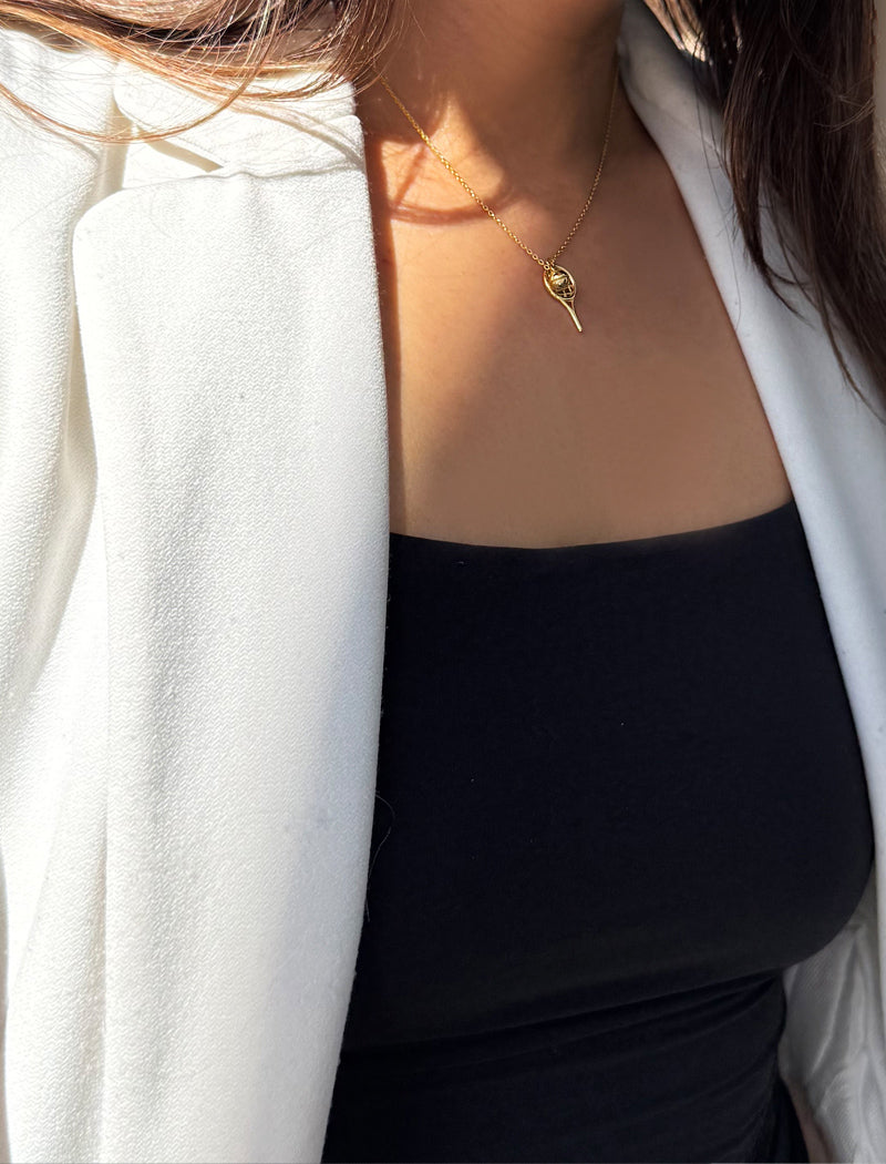 A close-up lifestyle shot of a model wearing a delicate gold tennis ball racquet necklace, styled with a black top and a white blazer. Designed by Sonia Hou, a celebrity AAPI inclusive demi-fine jewelry designer. Available in 18K Gold Over Sterling Silver, Sterling Silver and 18K Gold PVD Sterling Silver.