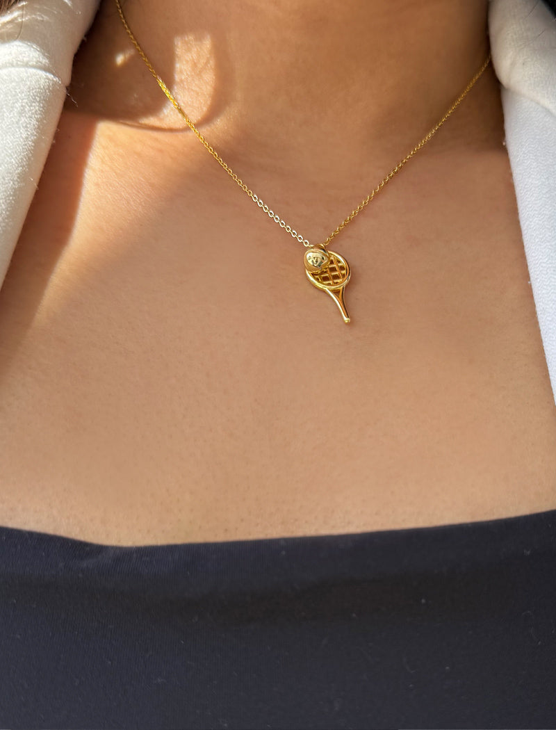 A close-up of a person's neck and chest wearing a delicate gold tennis racket pendant necklace with a tennis ball detail, styled with a white collared shirt and a dark navy top. Designed by Sonia Hou, a celebrity AAPI inclusive demi-fine jewelry designer. Available in 18K Gold Over Sterling Silver, Sterling Silver and 18K Gold PVD Sterling Silver.