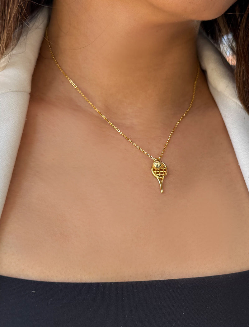 A close-up of a model wearing a delicate gold tennis ball racquet necklace, styled with a white blazer. Designed by Sonia Hou, a celebrity AAPI inclusive demi-fine jewelry designer. Available in 18K Gold Over Sterling Silver, Sterling Silver and 18K Gold PVD Sterling Silver (Waterproof).