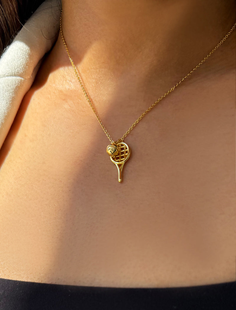 A close-up of a person's neck and chest wearing a delicate gold tennis racket pendant necklace with a tennis ball detail, set against warm sunlight and shadow. Designed by Sonia Hou, a celebrity AAPI inclusive demi-fine jewelry designer. Available in 18K Gold Over Sterling Silver, Sterling Silver and 18K Gold PVD Sterling Silver.