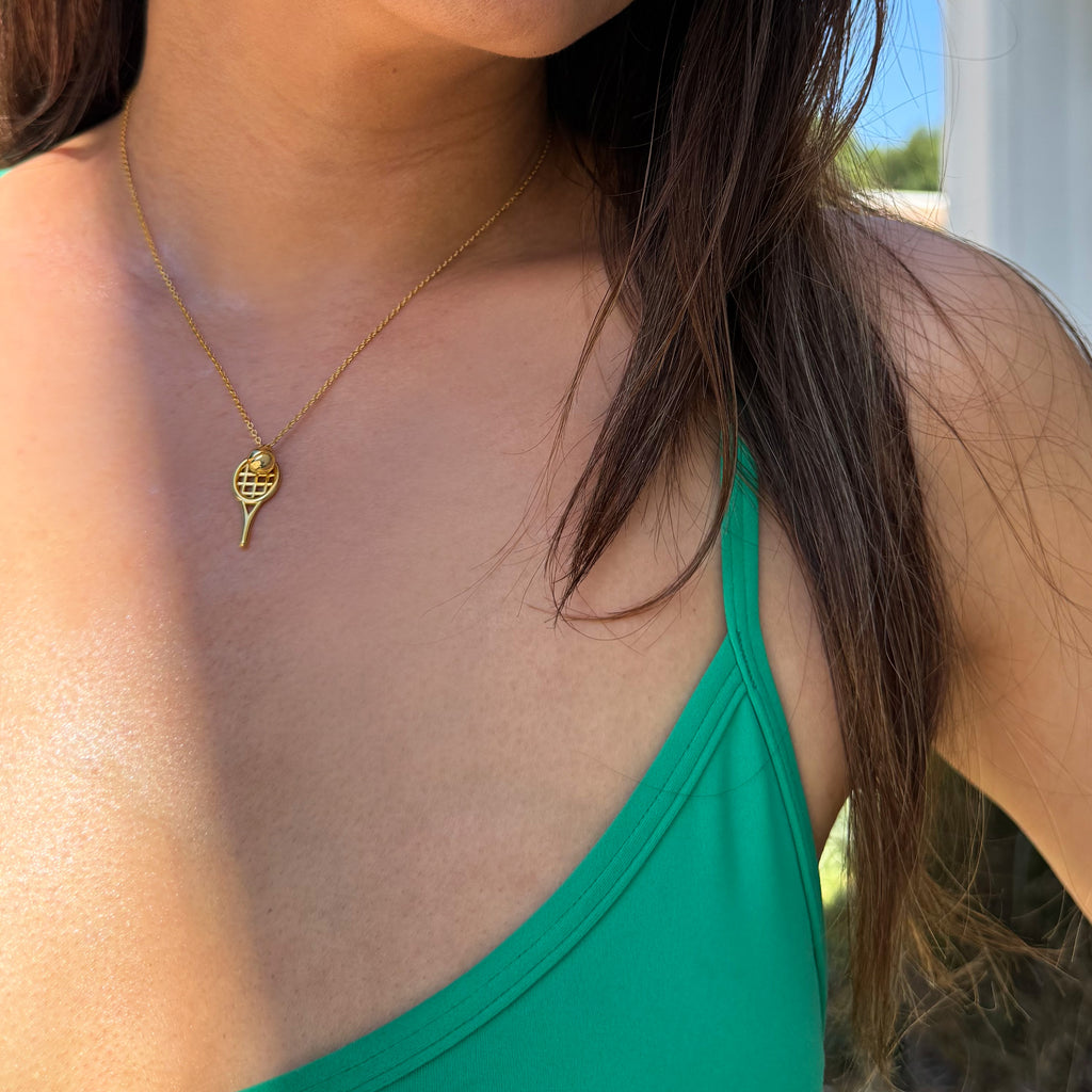 A close-up lifestyle shot of a woman wearing a delicate gold tennis ball and racket pendant necklace over a bright green tank top. Designed by Sonia Hou, a celebrity AAPI inclusive demi-fine jewelry designer. Available in 18K Gold Over Sterling Silver, Sterling Silver and 18K Gold PVD Sterling Silver (Waterproof).