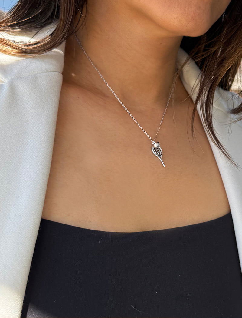 A close-up of a person wearing a delicate silver tennis racquet ball chain necklace with a small tennis ball pendant, styled with a white blazer and black top. Designed by Sonia Hou, a celebrity AAPI inclusive demi-fine jewelry designer. Available in 18K Gold Over Sterling Silver, Sterling Silver and 18K Gold PVD Sterling Silver (Waterproof).