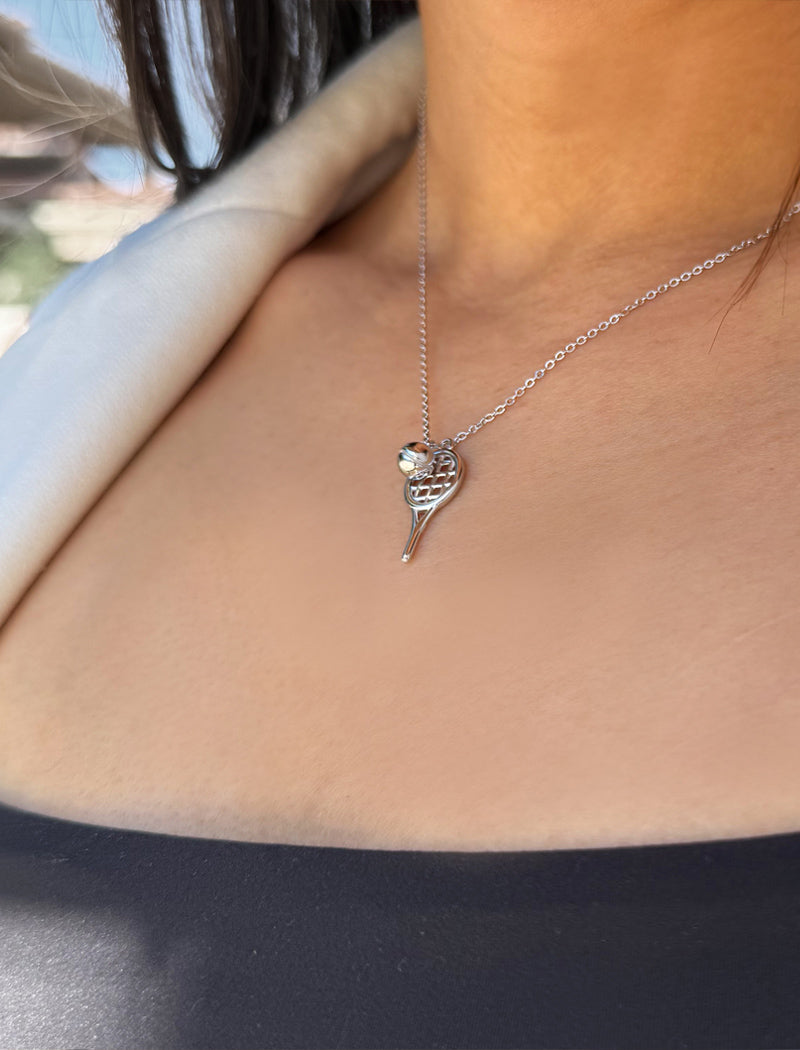 A close-up of a person wearing a delicate silver tennis racquet ball necklace featuring a small tennis ball, styled with a light-colored blazer. Designed by Sonia Hou, a celebrity AAPI inclusive demi-fine jewelry designer. Available in 18K Gold Over Sterling Silver, Sterling Silver and 18K Gold PVD Sterling Silver (Waterproof).
