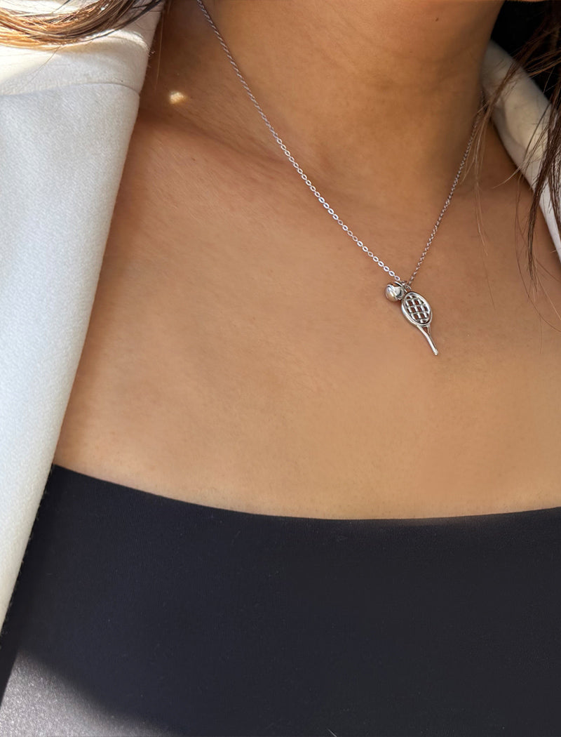 A close-up of a person's neck and chest wearing a delicate silver chain necklace with a tennis racket pendant and a tennis ball. Designed by Sonia Hou, a celebrity AAPI inclusive demi-fine jewelry designer. Available in 18K Gold Over Sterling Silver, Sterling Silver and 18K Gold PVD Sterling Silver (Waterproof).