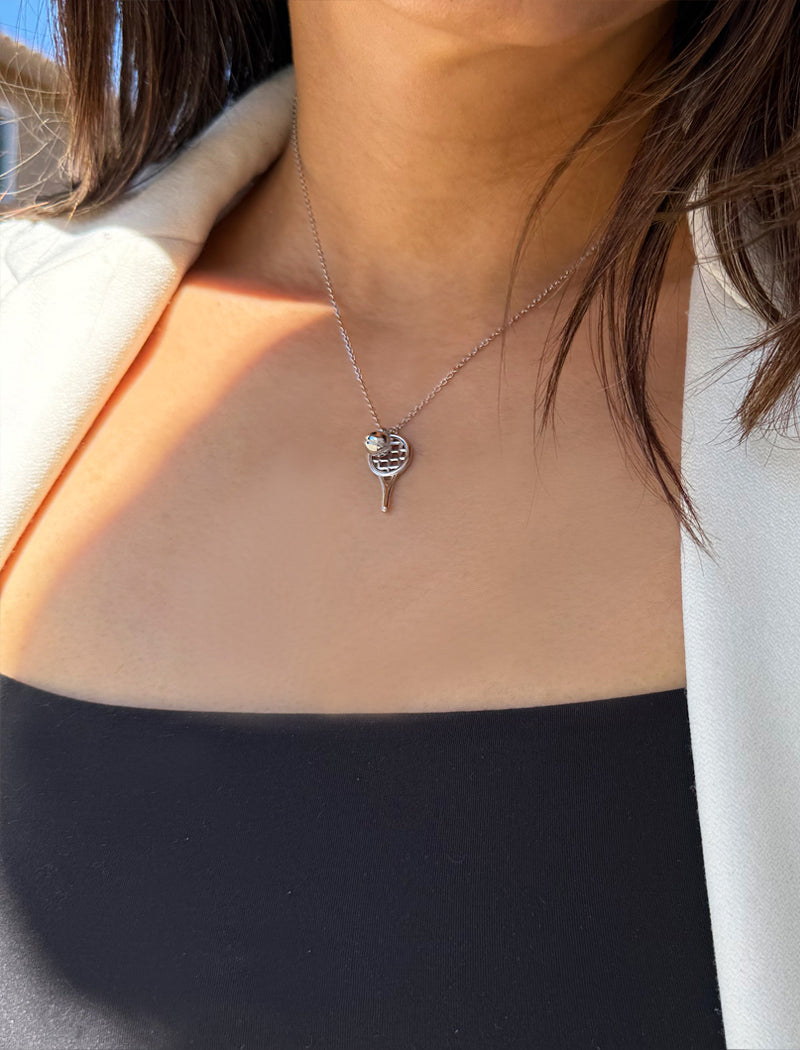 A close-up of a person wearing a delicate silver tennis racquet ball necklace featuring a tiny dangling tennis ball, styled with a black top and a light beige blazer. Designed by Sonia Hou, a celebrity AAPI inclusive demi-fine jewelry designer. Available in 18K Gold Over Sterling Silver, Sterling Silver and 18K Gold PVD Sterling Silver (Waterproof).